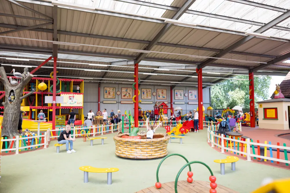 Ein farbenfrohes Indoor-Spielparadies in einer großen, überdachten Halle, der von vielen Familien genutzt wird. Im Vordergrund sitzt eine Frau links auf einer Bank, daneben steht ein künstlicher Baumstamm mit Vögeln. Ein zentrales Karussell in Weidenkorb-Optik mit einer sitzenden Person steht auf der grünen Gummibodenfläche. Links ragt ein großes mehrstöckiges Kletterlabyrinth empor. Farbige Zäune grenzen verschiedene Bereiche ab. An der Rückwand sind bunte Wandgemälde mit Kindermotiven zu sehen. Erwachsene und Kinder bewegen sich durch die Halle. Rote Säulen und die Stahlkonstruktion des Dachs sind sichtbar, im Hintergrund Bäume.