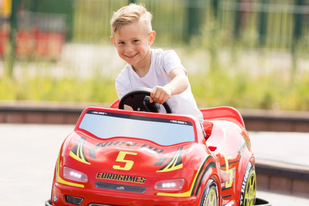 junge-in-potzi-kart Ein freudestrahlender kleiner Junge mit blonden Haaren sitzt am Steuer eines leuchtend roten Spielzeugautos (POTZI-Kart) mit der Startnummer 5 auf einer Außenanlage.